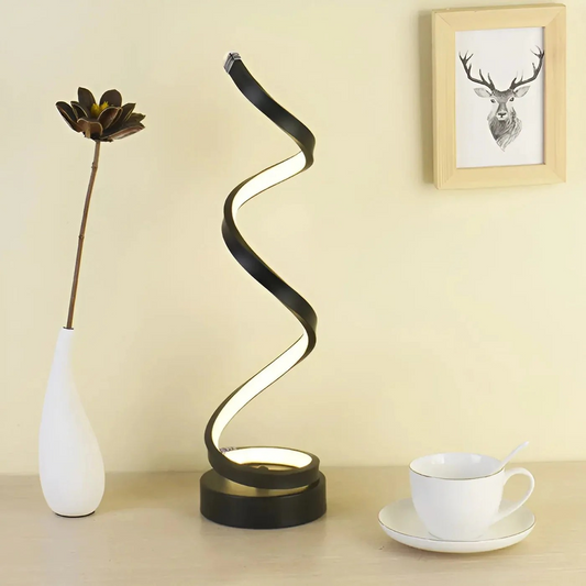Modern table lamp with a curved design on a desk next to a vase and cup.